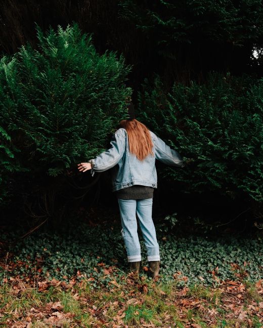 Full body back view of unrecognizable female in casual clothes putting head in foliage of shrub