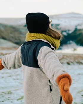 A woman in winter clothing extends her arms in a snowy outdoor setting, embracing the cold weather.