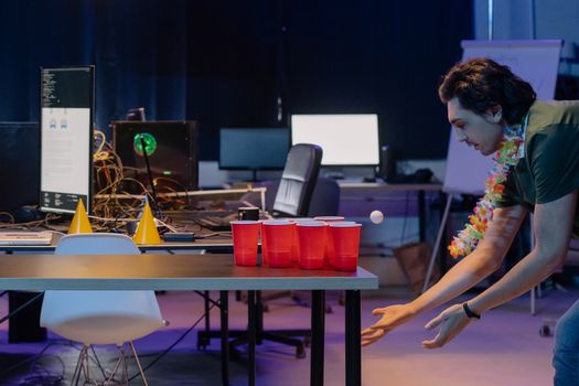 A fun beer pong game in an office setting with a man throwing a ping pong ball towards red cups.