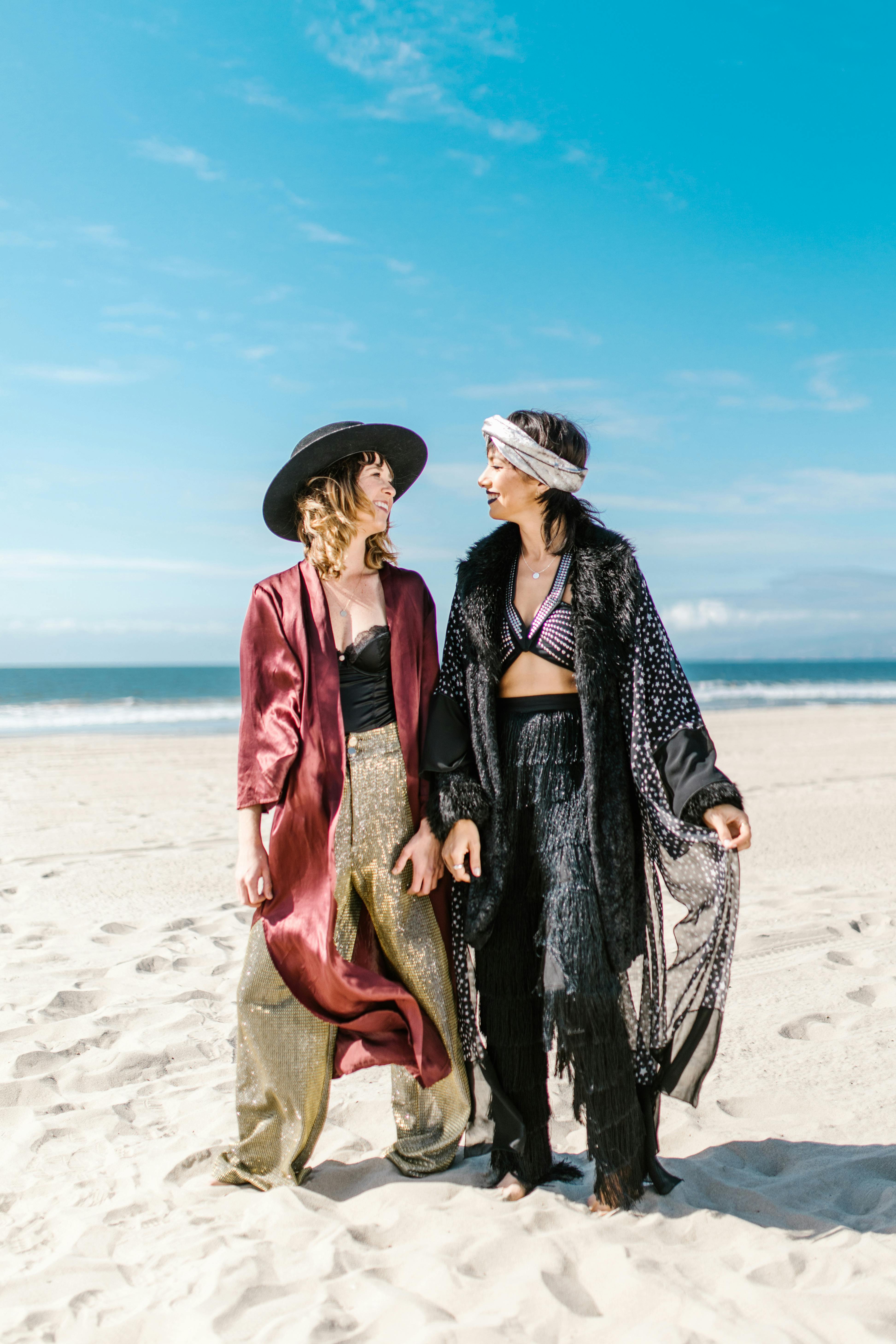 Two women in bohemian attire enjoy a sunny day at the beach, embracing a carefree vibe.