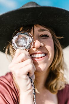 A smiling woman with a hat holds a prism crystal outdoors in sunlight.