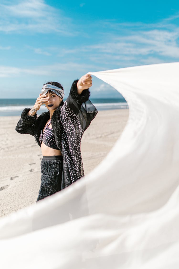 A Woman In Black Outfit Swaying The White Fabric She Is Holding While Standing On The Shore Of The Beach