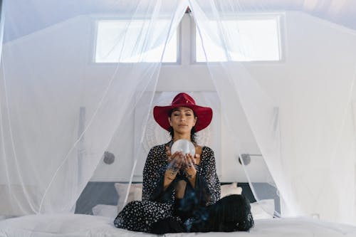 Woman in Red Hat and Black Outfit Sitting on the Bed while Holding a Crystal Ball