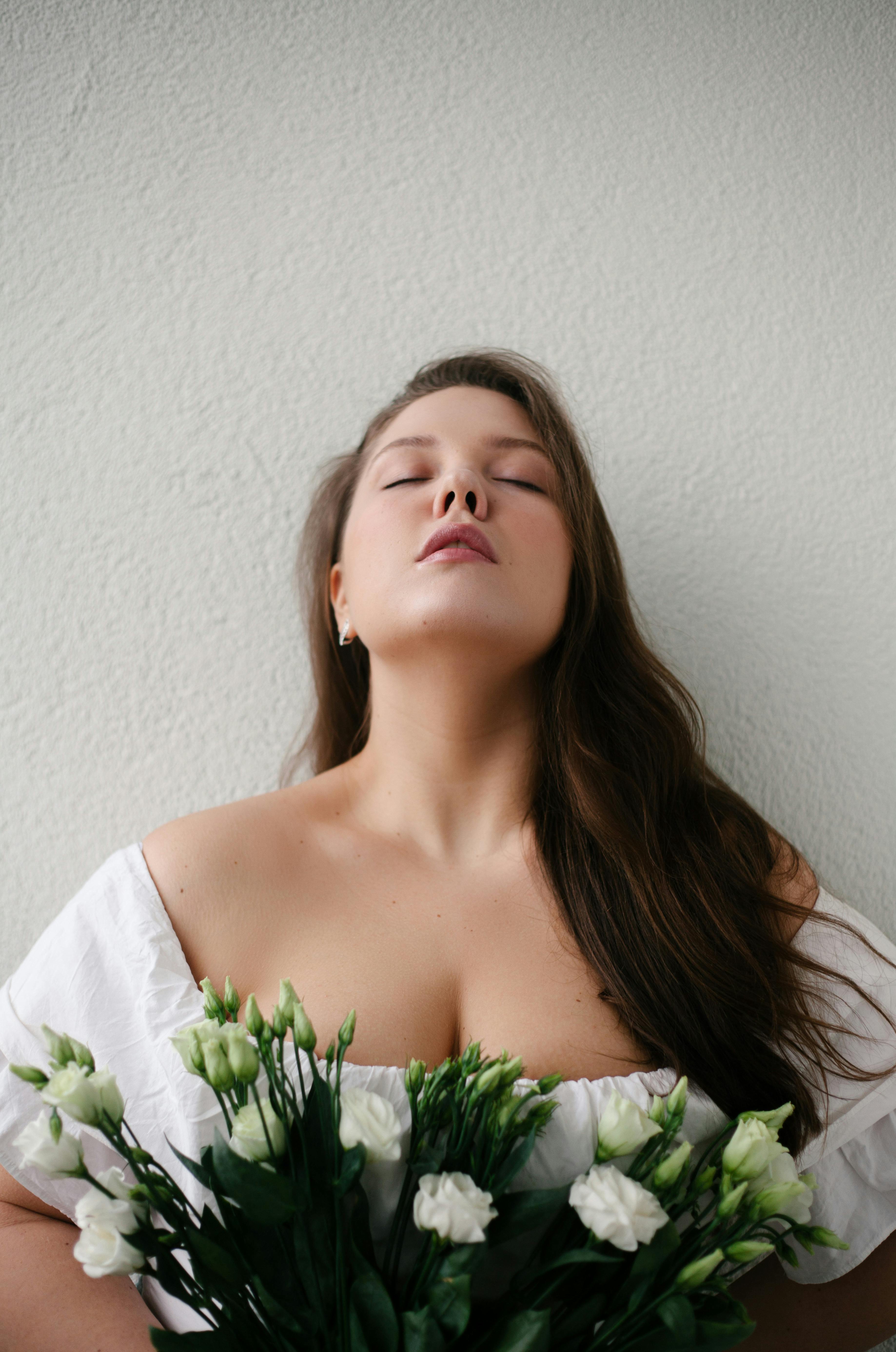 Thoughtful young lady in white blouse with closed eyes near wall with blooming bouquet of white roses in light room