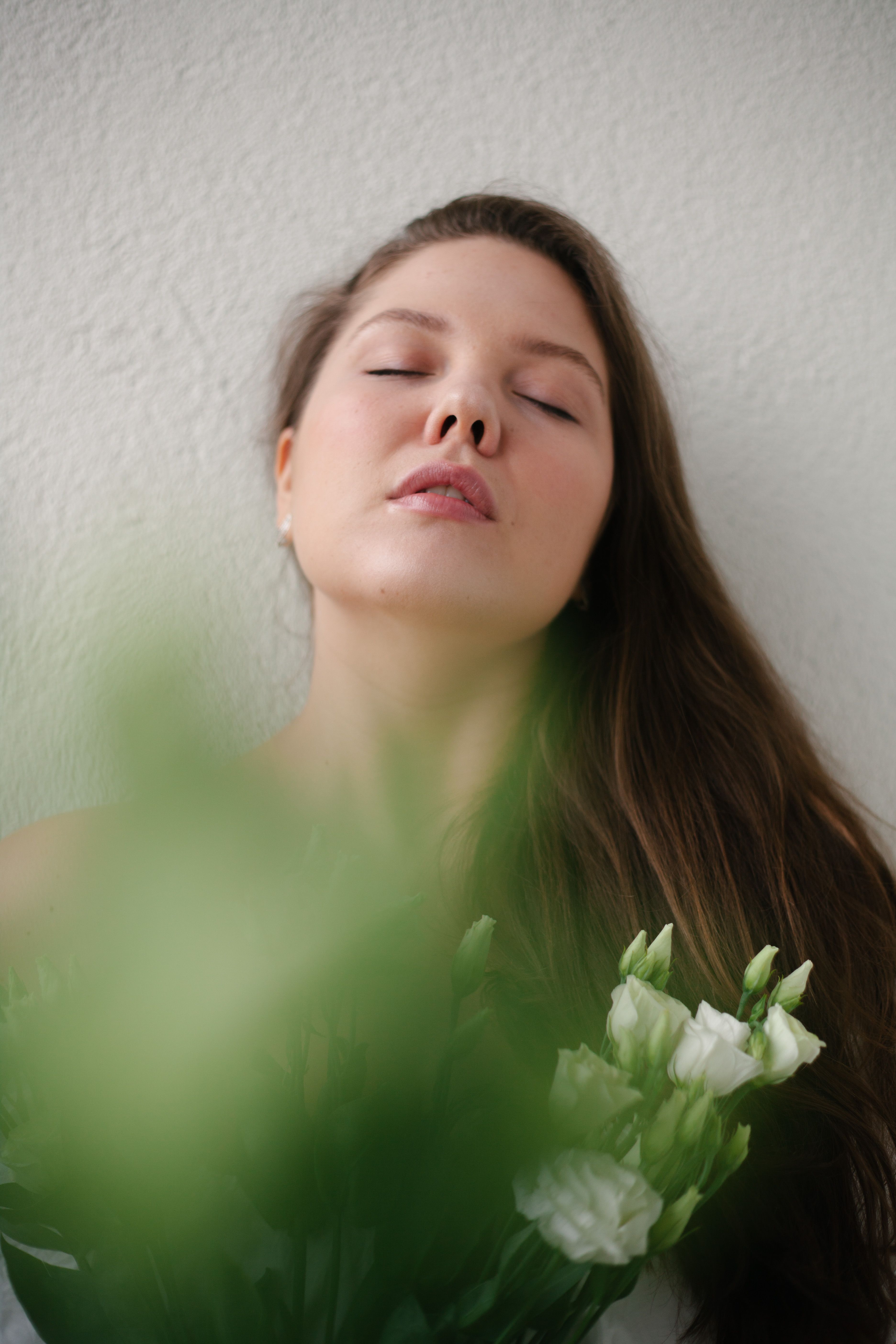 Dreamy young lady in white blouse with closed eyes standing near wall with blooming bouquet of white roses in hands in light place