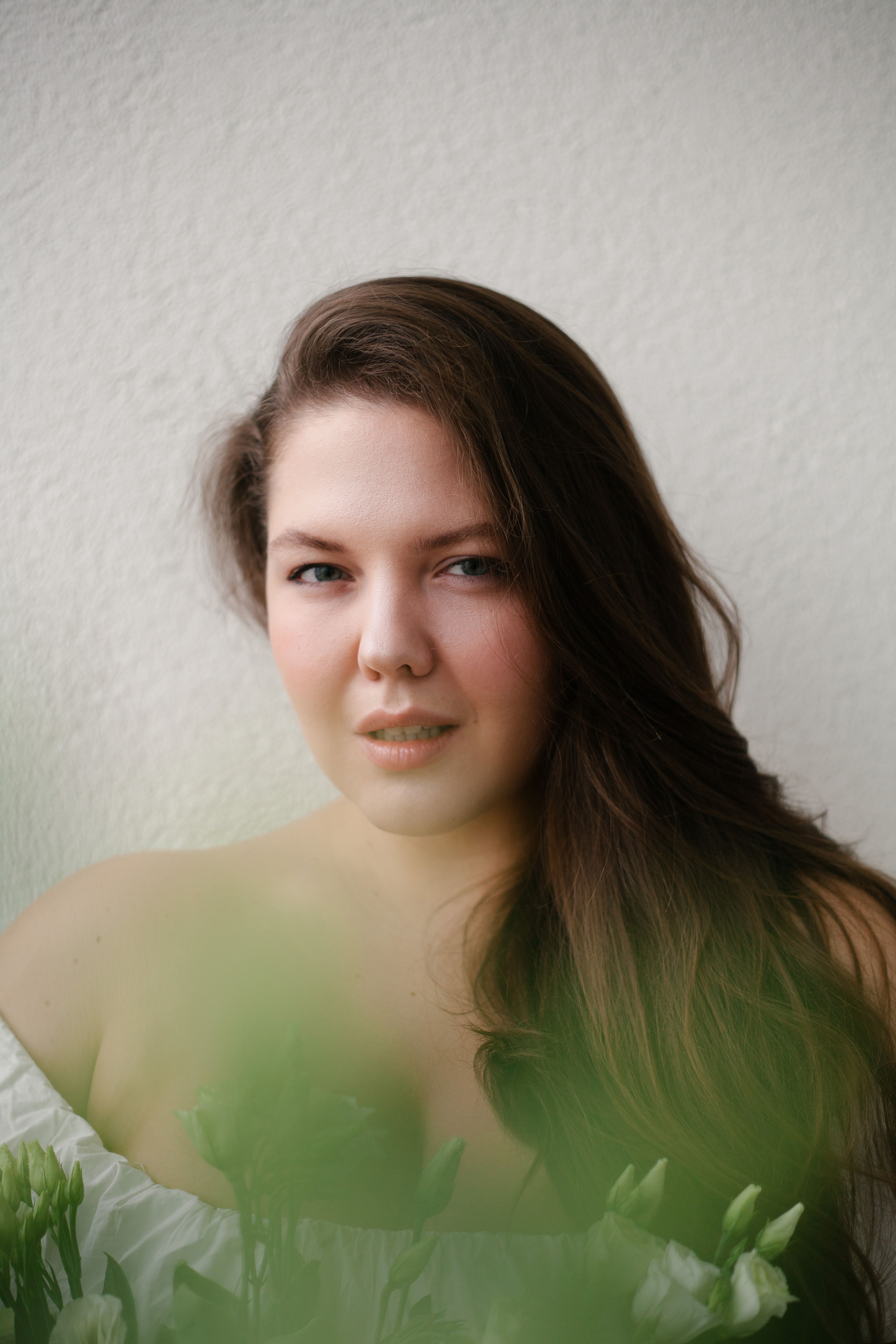 Pensive young female in white blouse with blossoming bouquet of white flowers near wall looking at camera in bright room