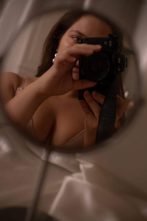 Anonymous lady in bra with photo camera taking selfie in reflection of cosmetic mirror in light room