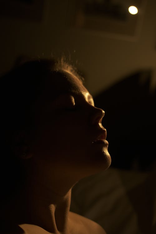 Young woman with bare shoulders and closed eyes in dark room with sunlight on body