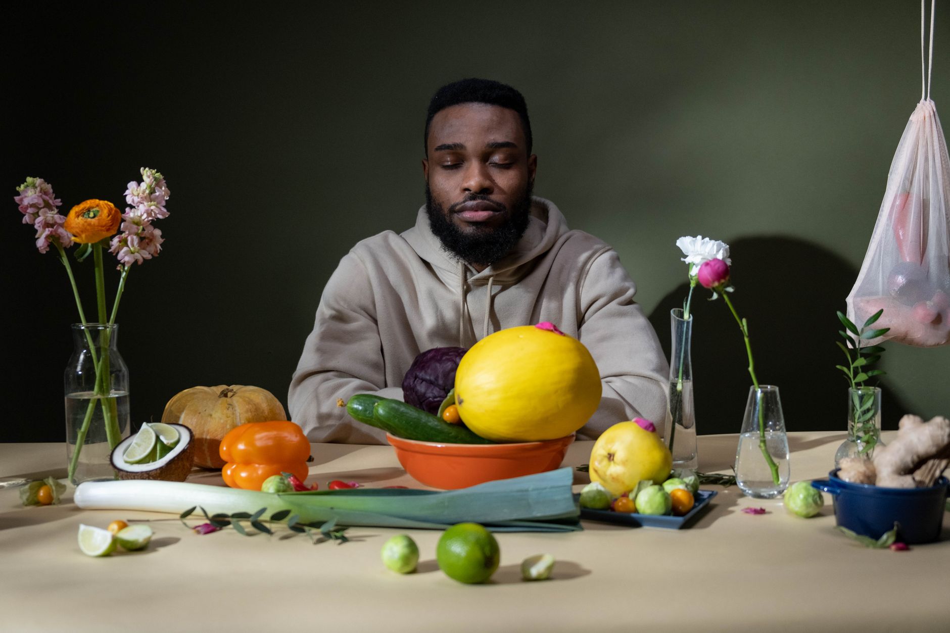 Man in hoodie meditates among fresh vegetables and flowers on a table.
