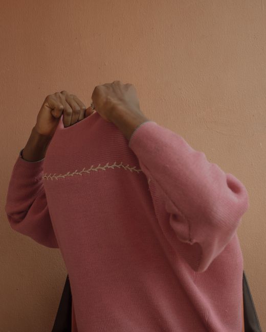 An individual in a pink sweater adjusts the garment, blending into a warm neutral background.