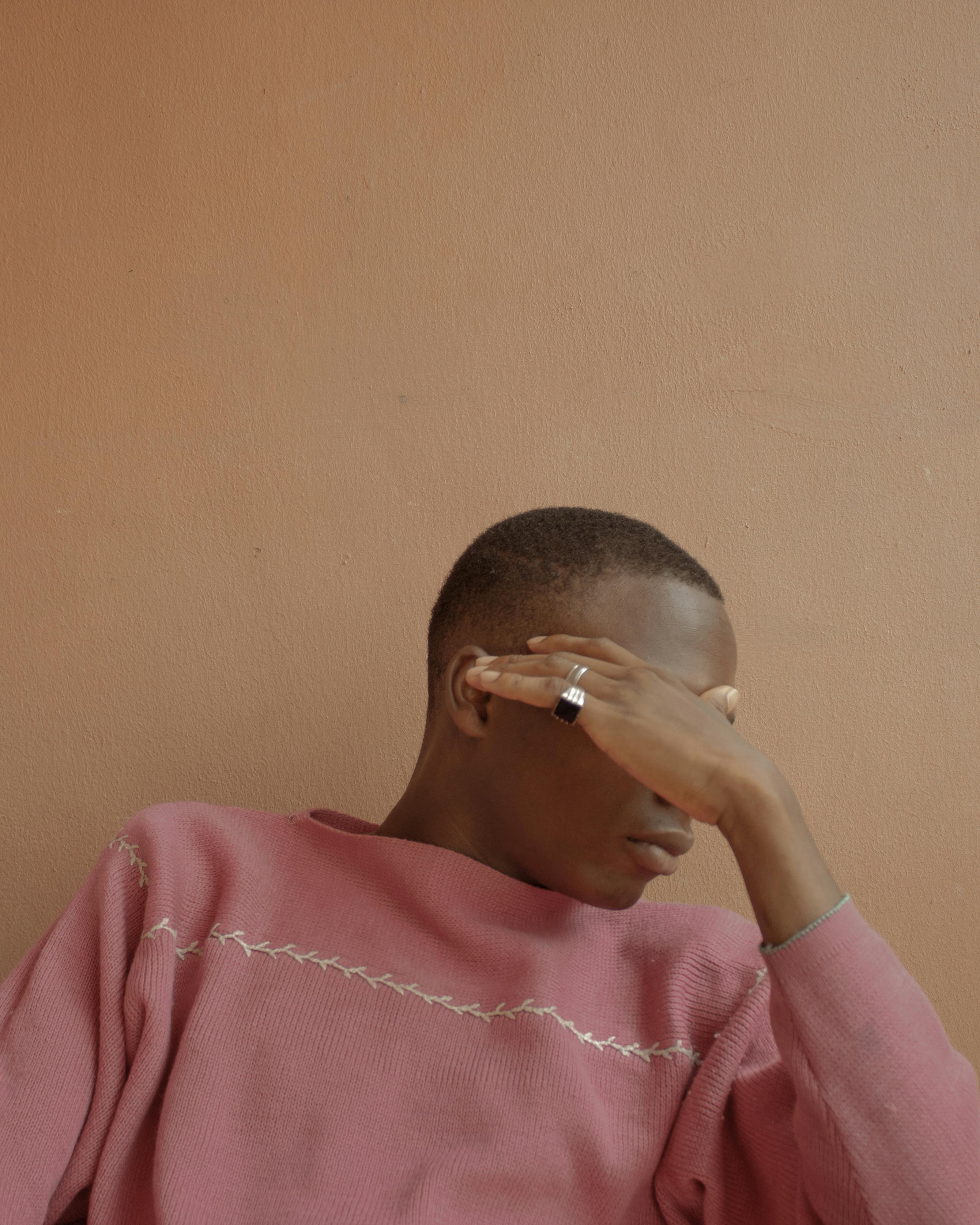 Young man covering face with hand while seated, wearing a pink sweater against a peach wall.