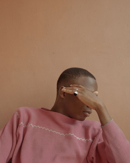 Young man covering face with hand while seated, wearing a pink sweater against a peach wall.