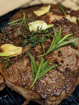 Close-up of a succulent steak topped with fresh herbs and garlic, sizzling on the grill.
