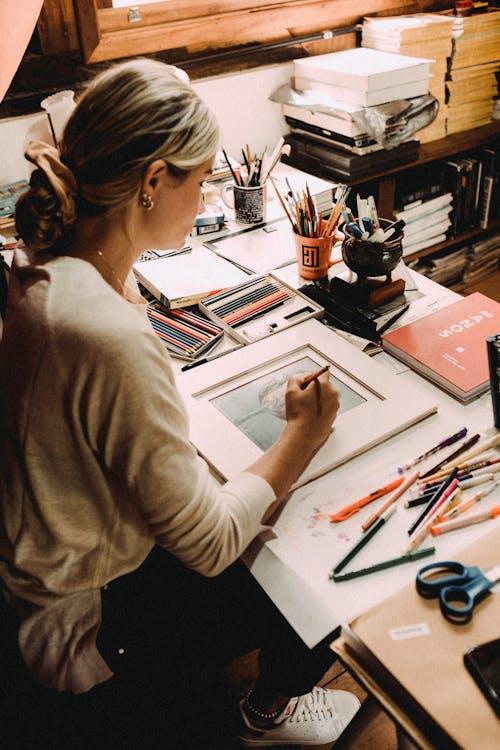 Free From above of young concentrated woman creating picture at table with many art supplies Stock Photo