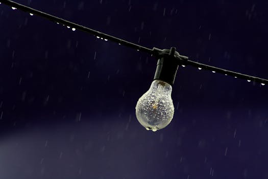 A clear lightbulb with raindrops hanging on a wire against a deep blue sky.
