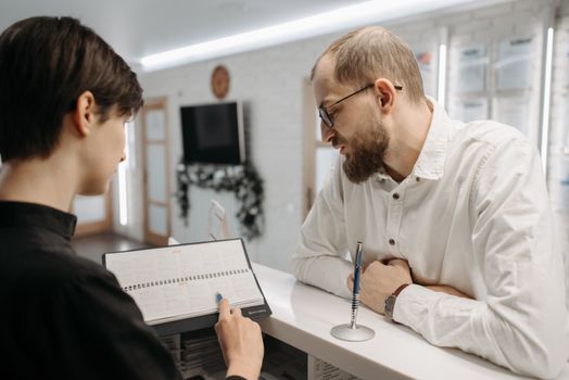 A receptionist and client converse over an appointment book at a clinic reception desk.