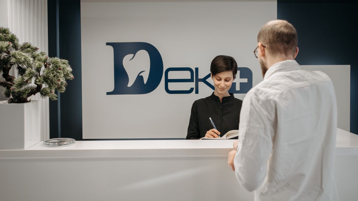 A receptionist assisting a patient at a dental clinic counter, with a potted plant in sight.
