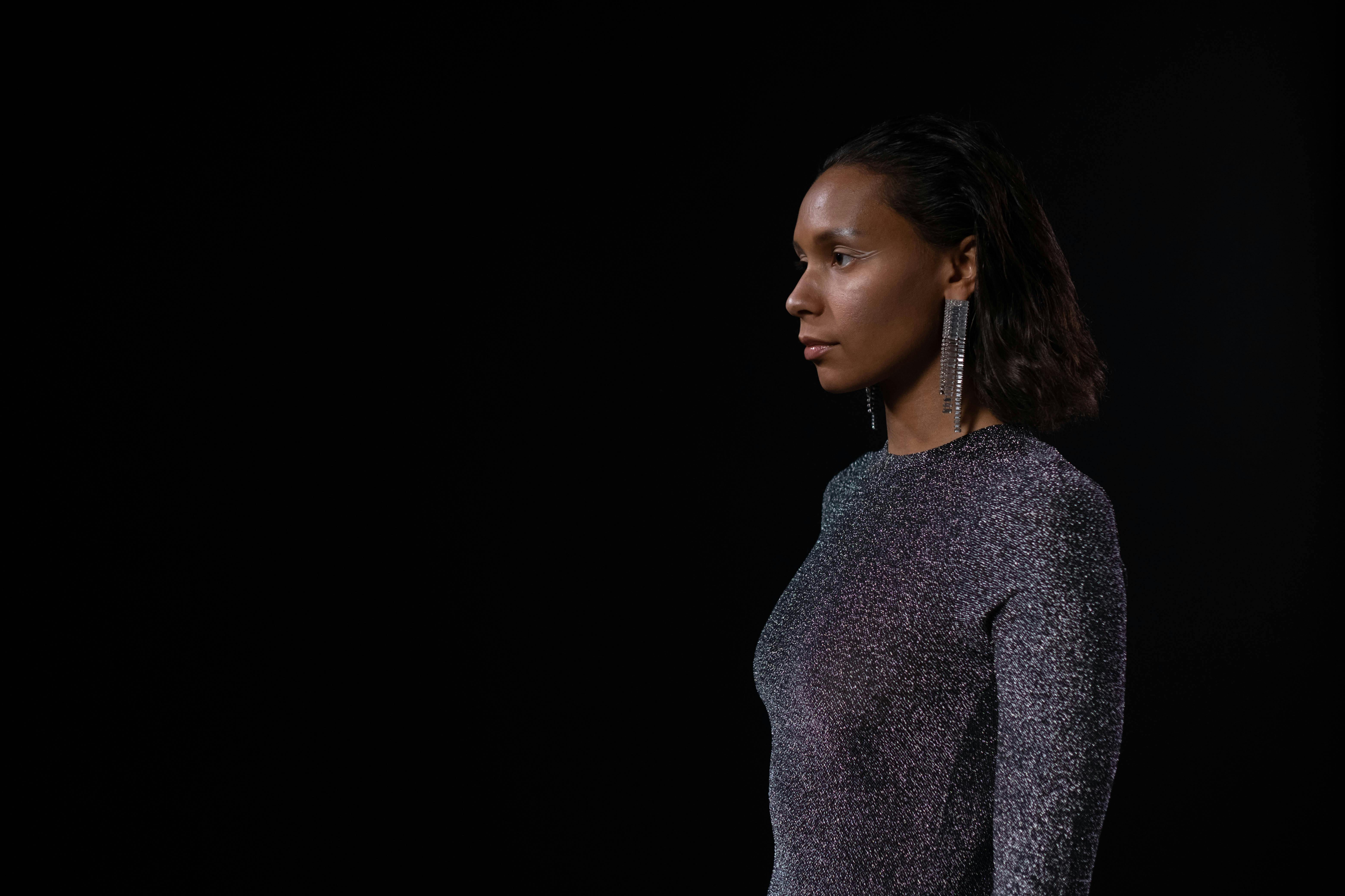 Profile view of a poised woman in a sparkling gray top with a dramatic earring, set against a black background.