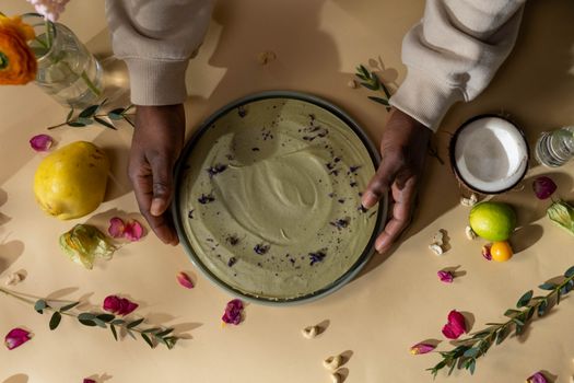 A top-down view of a vegan matcha cake with vibrant floral and fruit decorations, held by hands.