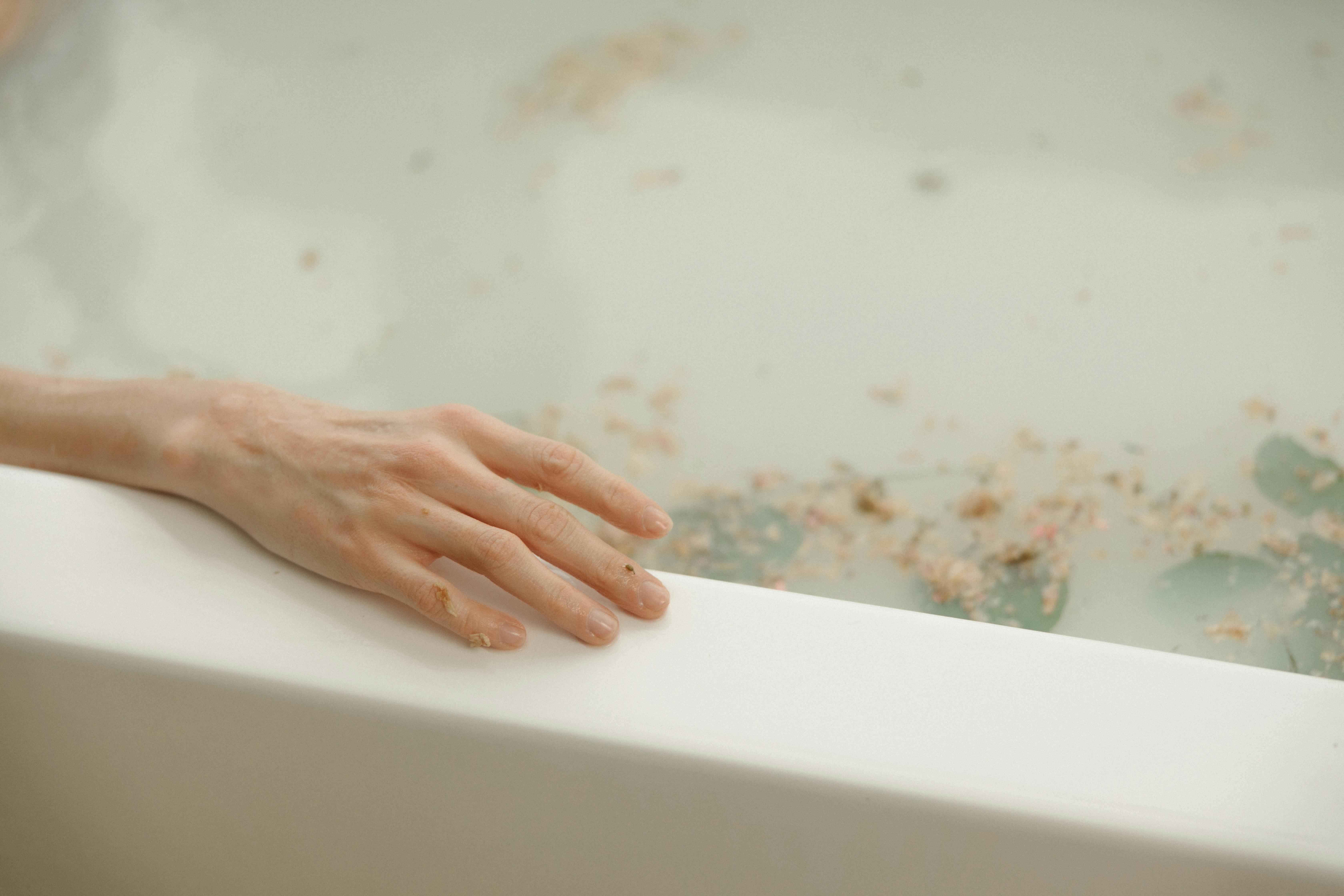 A serene hand resting on a bathtub rim surrounded by floating petals.