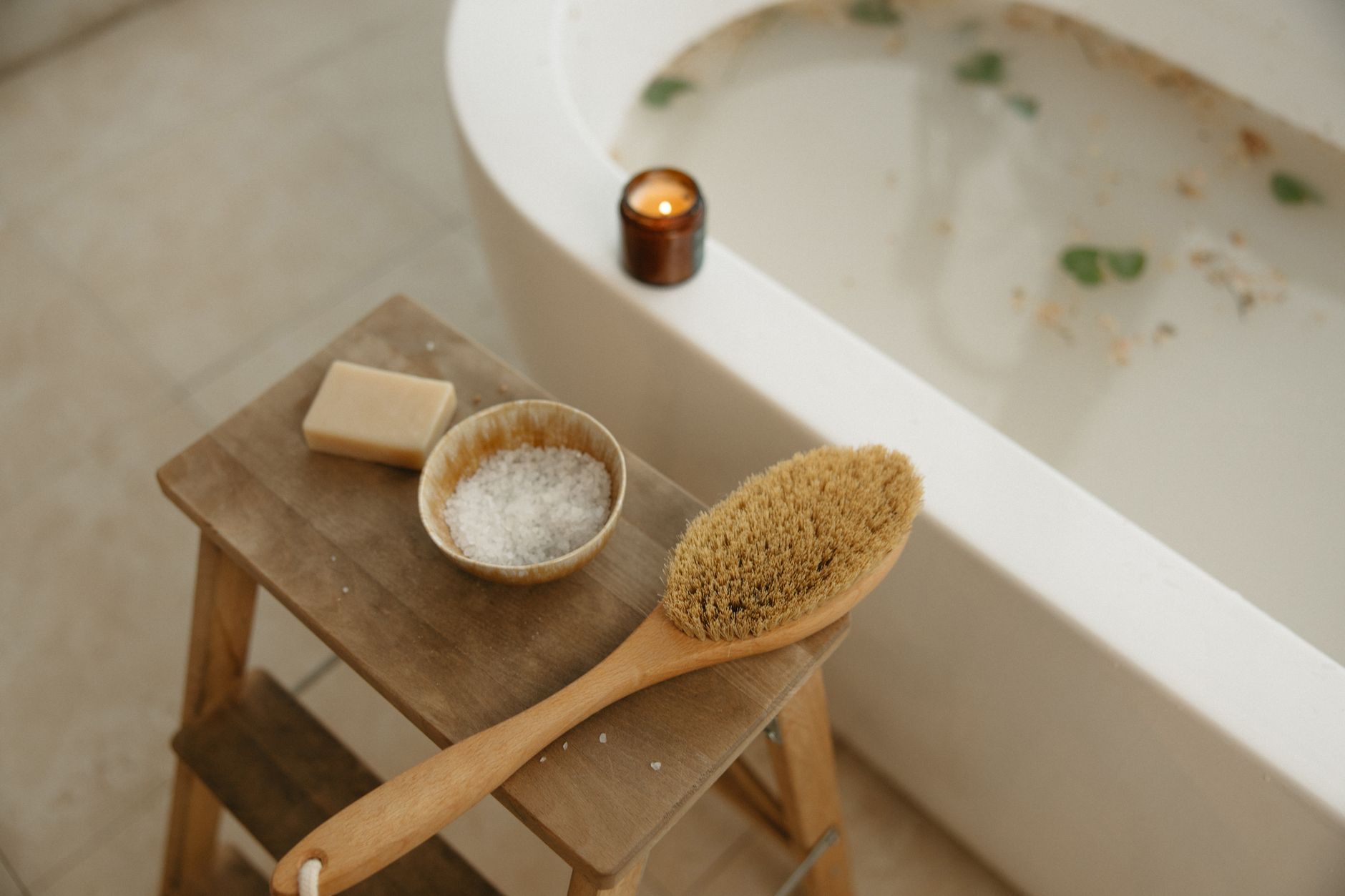 epsom salt detox bath — Relaxation bath scene with candle, dry brush, sea salt, and soap on a wooden stool for a serene home spa experience.