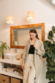A woman stands in a modern bathroom with plants and stylish decor.
