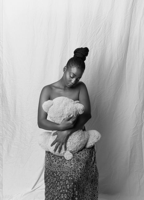 Black and white of serious young black lady in skirt with bare shoulders hugging soft bear toy while sitting with sheet on background in studio