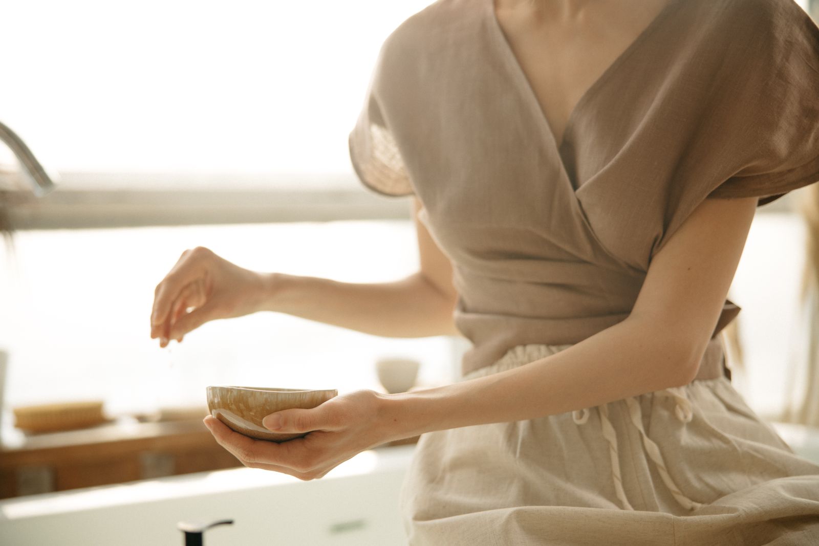 Woman in a taupe linen wrap top holding a small ceramic bowl in warm window light
