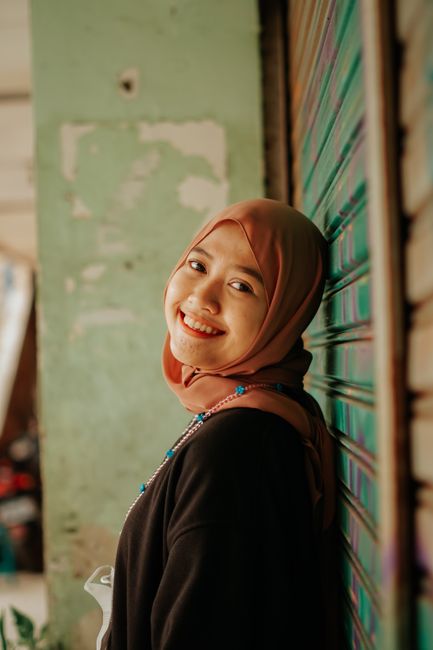 A cheerful woman in a hijab is smiling while leaning against a roll-up door in an outdoor setting.
