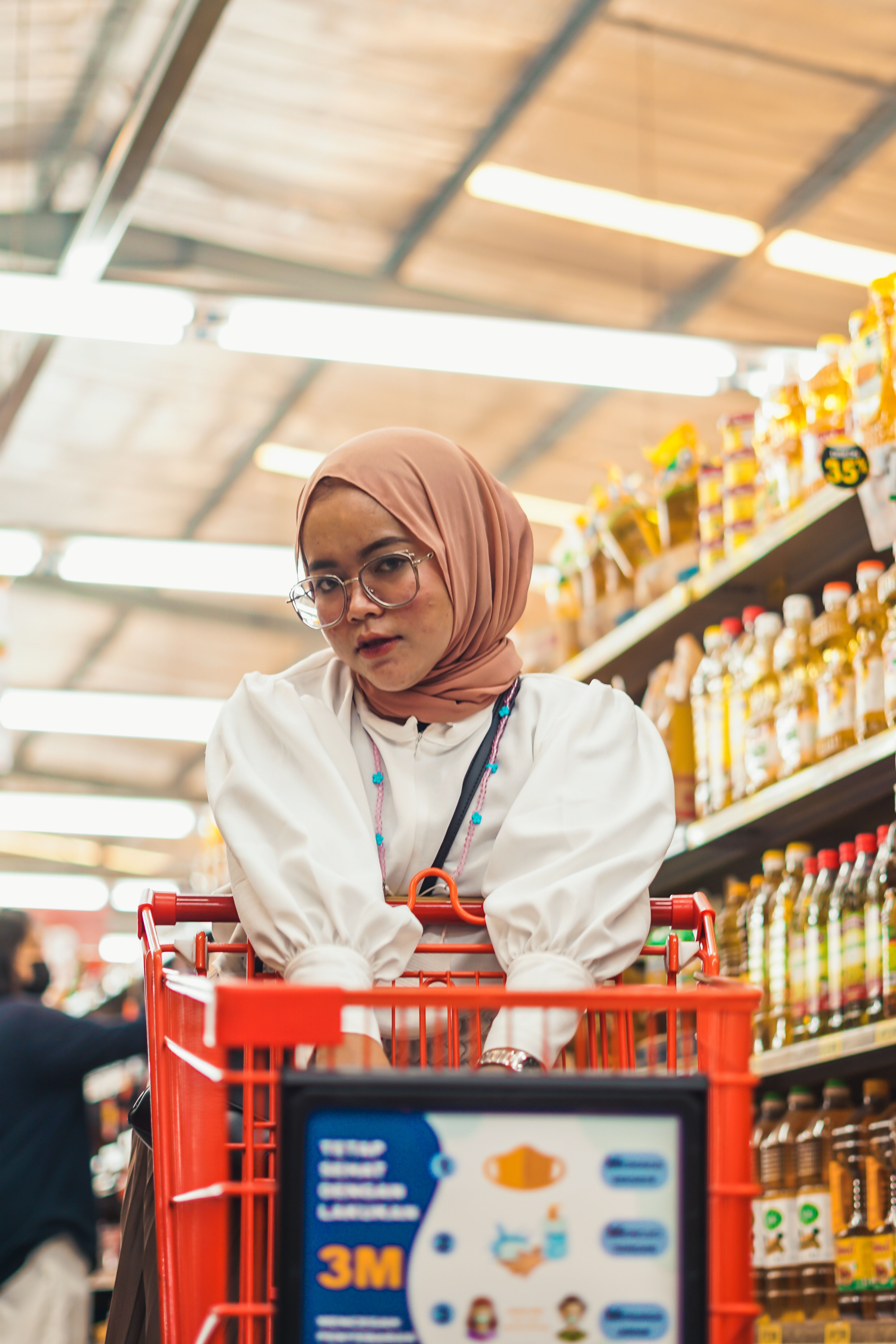 Asian woman in hijab with shopping cart in a grocery store aisle. Warm lighting and organized shelves create a cozy atmosphere.