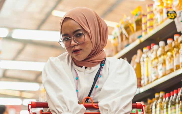Asian woman in hijab with shopping cart in a grocery store aisle. Warm lighting and organized shelves create a cozy atmosphere.