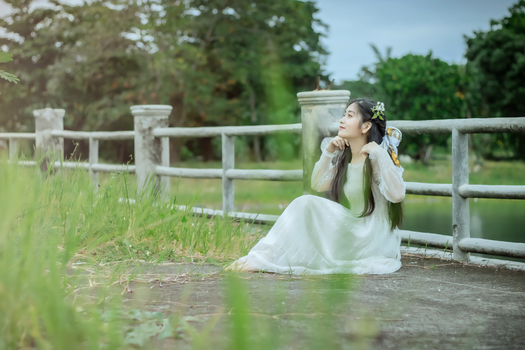 Asian woman in white dress seated by a fence in a lush green outdoor setting.