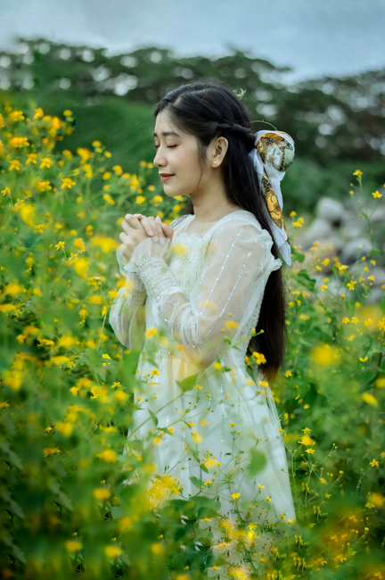 Woman in white dress with eyes closed, praying amidst vibrant yellow flowers.
