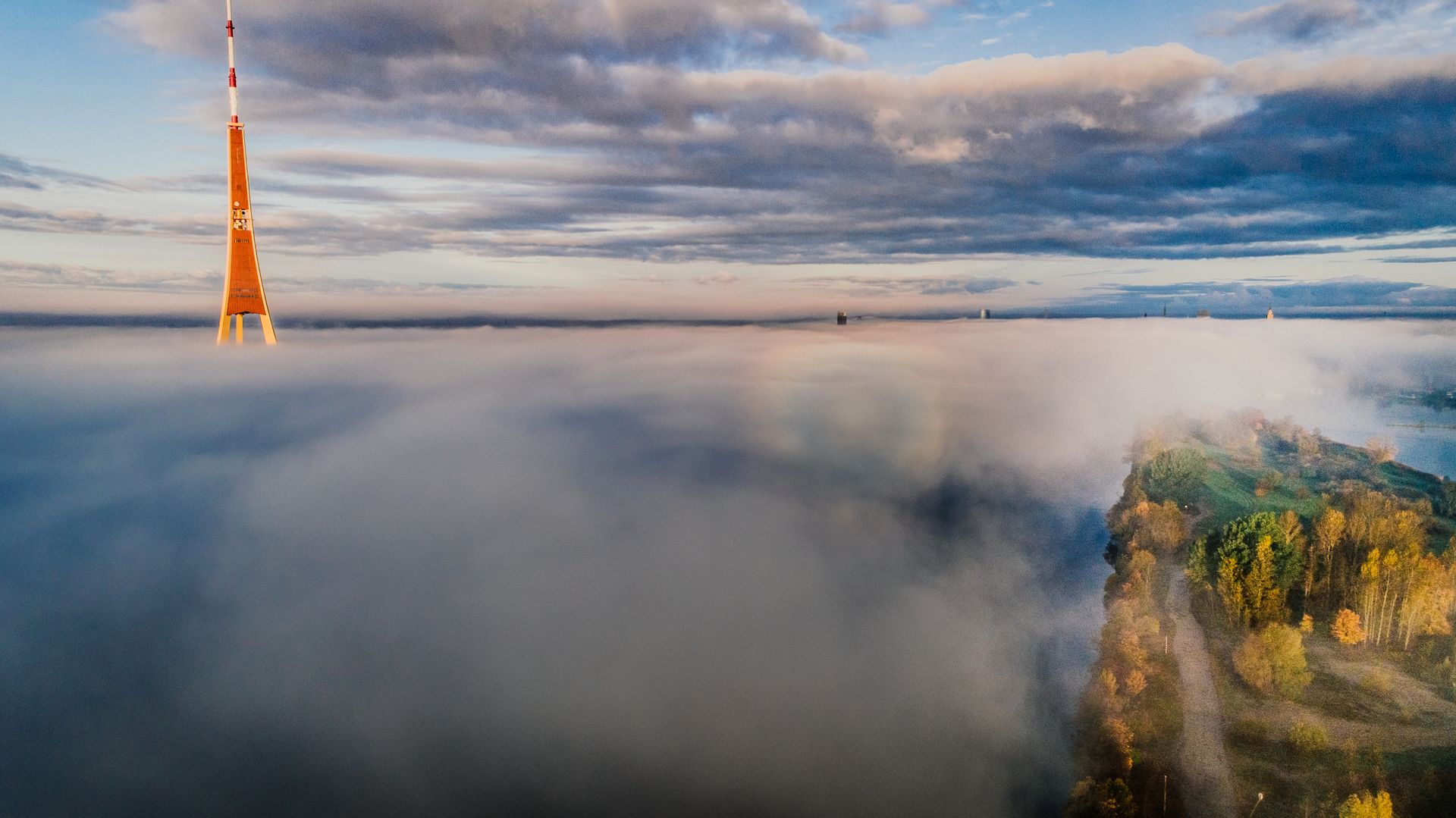 Aerial shot of a tower rising above clouds with a scenic landscape below.