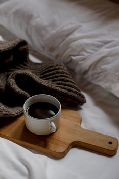A comforting scene featuring a cup of coffee on a wooden board next to a warm knitted sweater on a bed.
