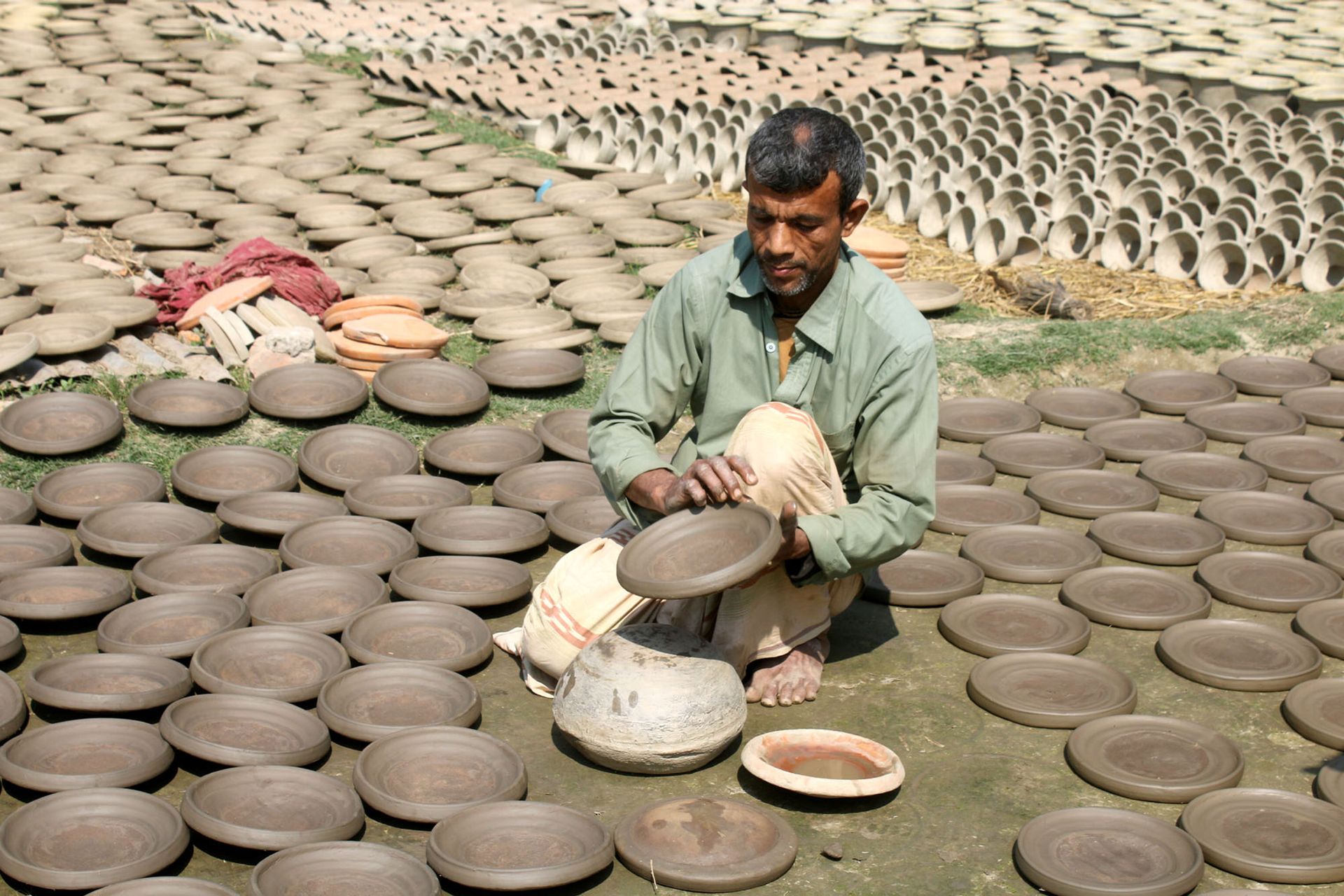 A dedicated artisan skillfully crafts pottery outdoors, surrounded by clay dishes drying in the sun.