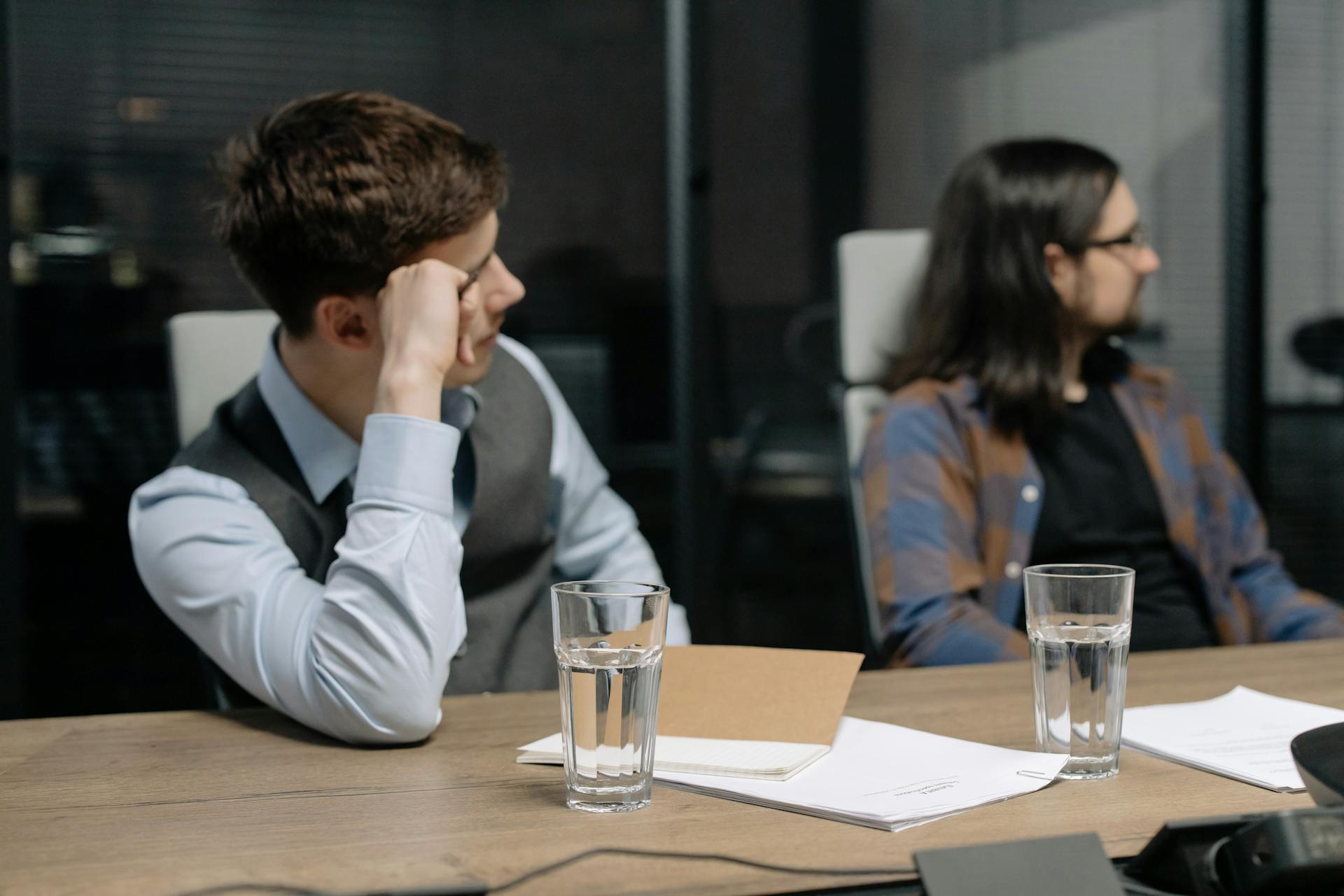 Two young adults in a business meeting setting with focus on written documents and water glasses.