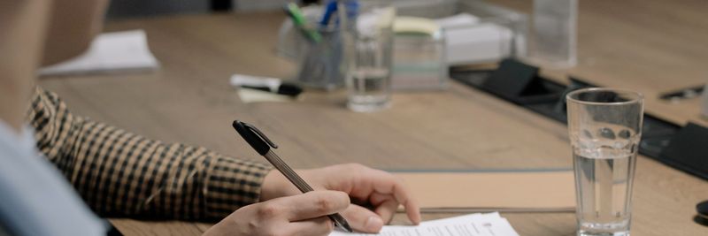 Person in business attire signing a document at a wooden table in an office setting.