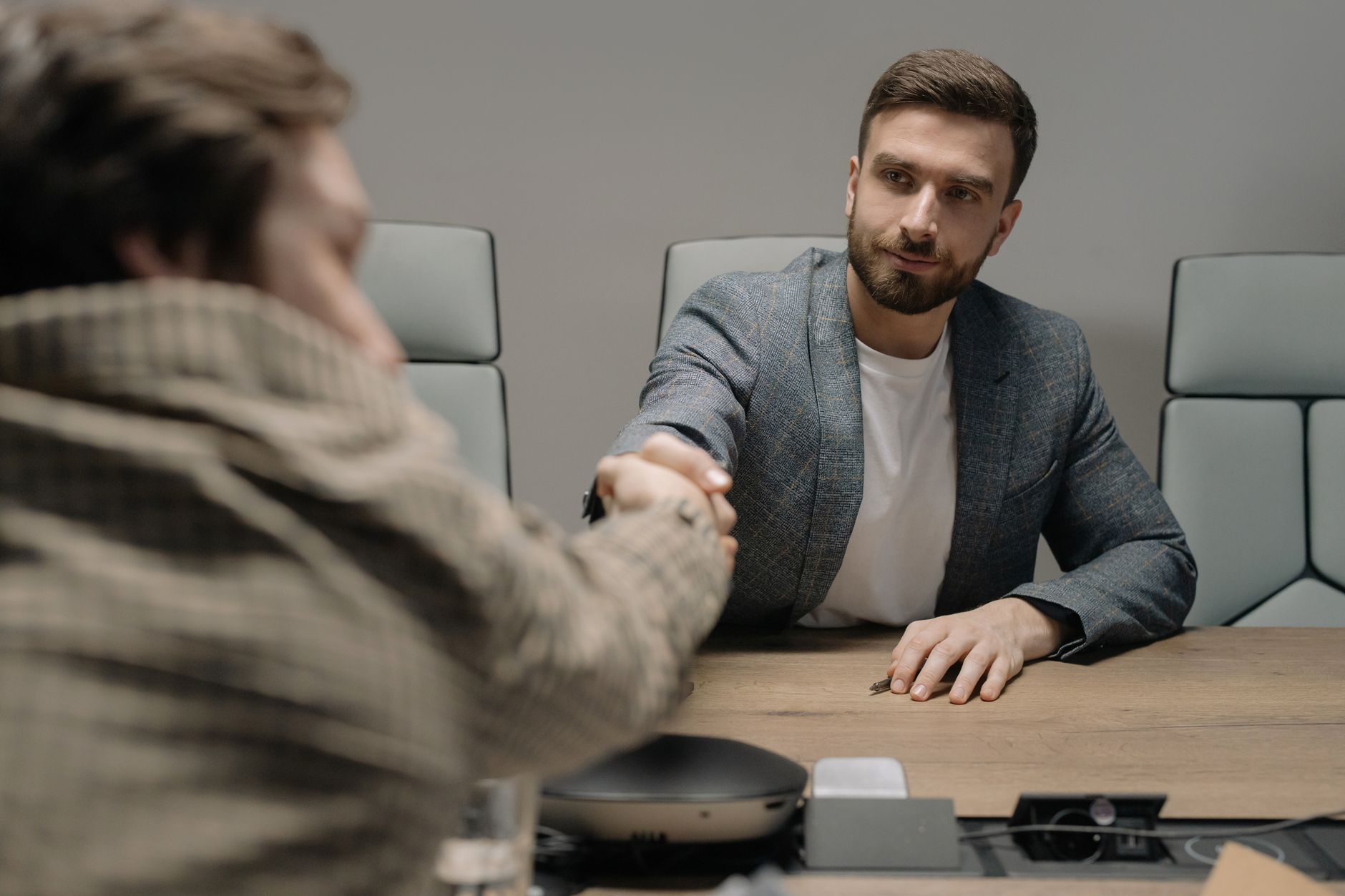 Two businessmen shaking hands across a wooden desk in a modern office setting.