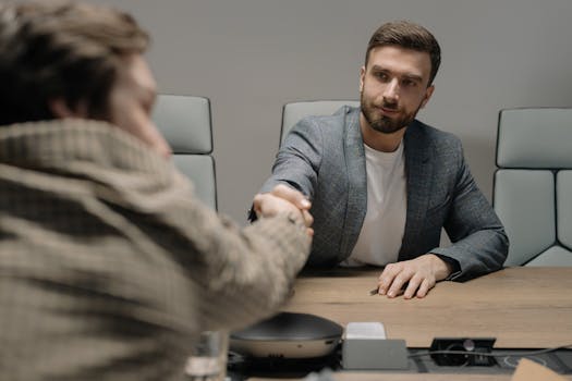 Two businessmen shaking hands across a wooden desk in a modern office setting.