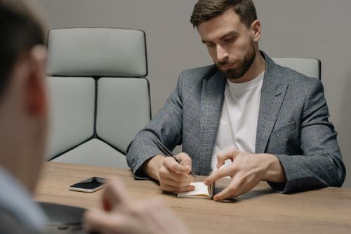 Un homme barbu en veste grise est assis à une table de réunion et prend des notes dans un petit carnet, tandis qu'un autre interlocuteur est visible au premier plan, flou.