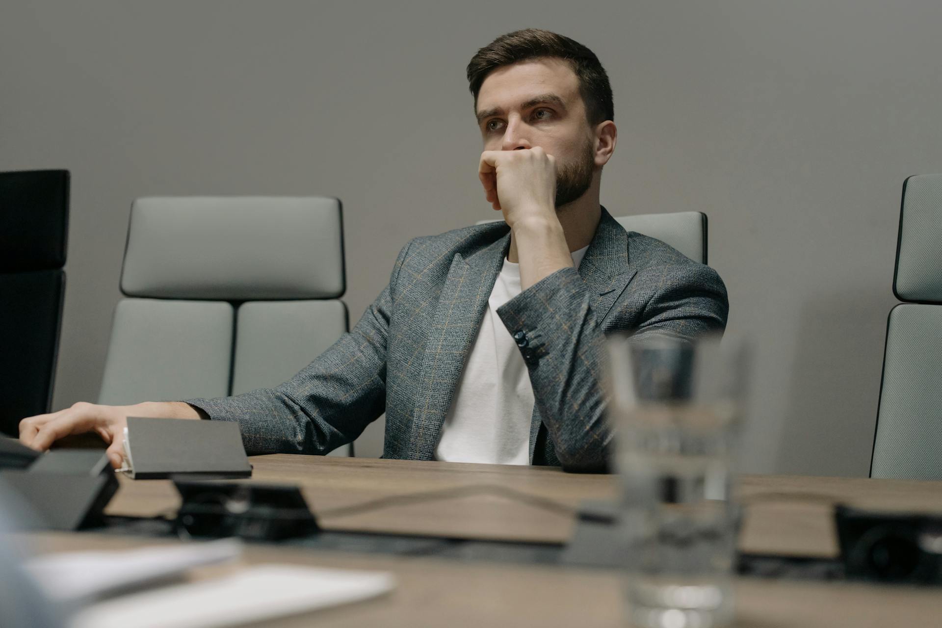 A contemplative businessman sitting at a meeting table in a modern office setting.