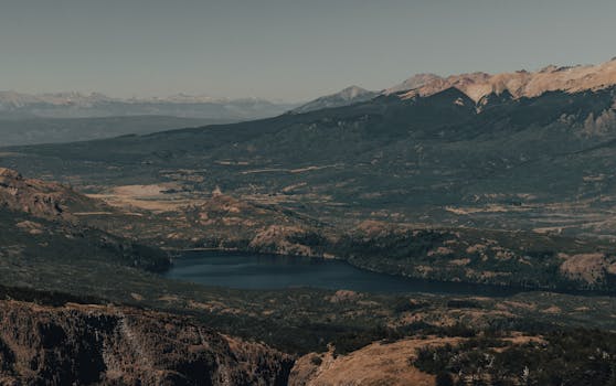 Areal view of mountainous terrain with green trees and plants near hills and pond under cloudless gray sky in daylight