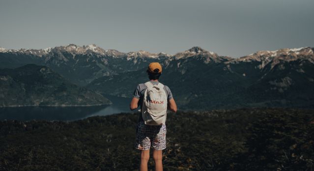 Back view of unrecognizable man in casual outfit with backpack standing on hill and admiring view of mountains near river and green plants with trees under cloudless sky in daytime