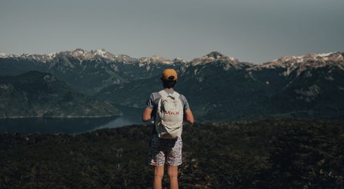 Free Back view of unrecognizable man in casual outfit with backpack standing on hill and admiring view of mountains near river and green plants with trees under cloudless sky in daytime Stock Photo