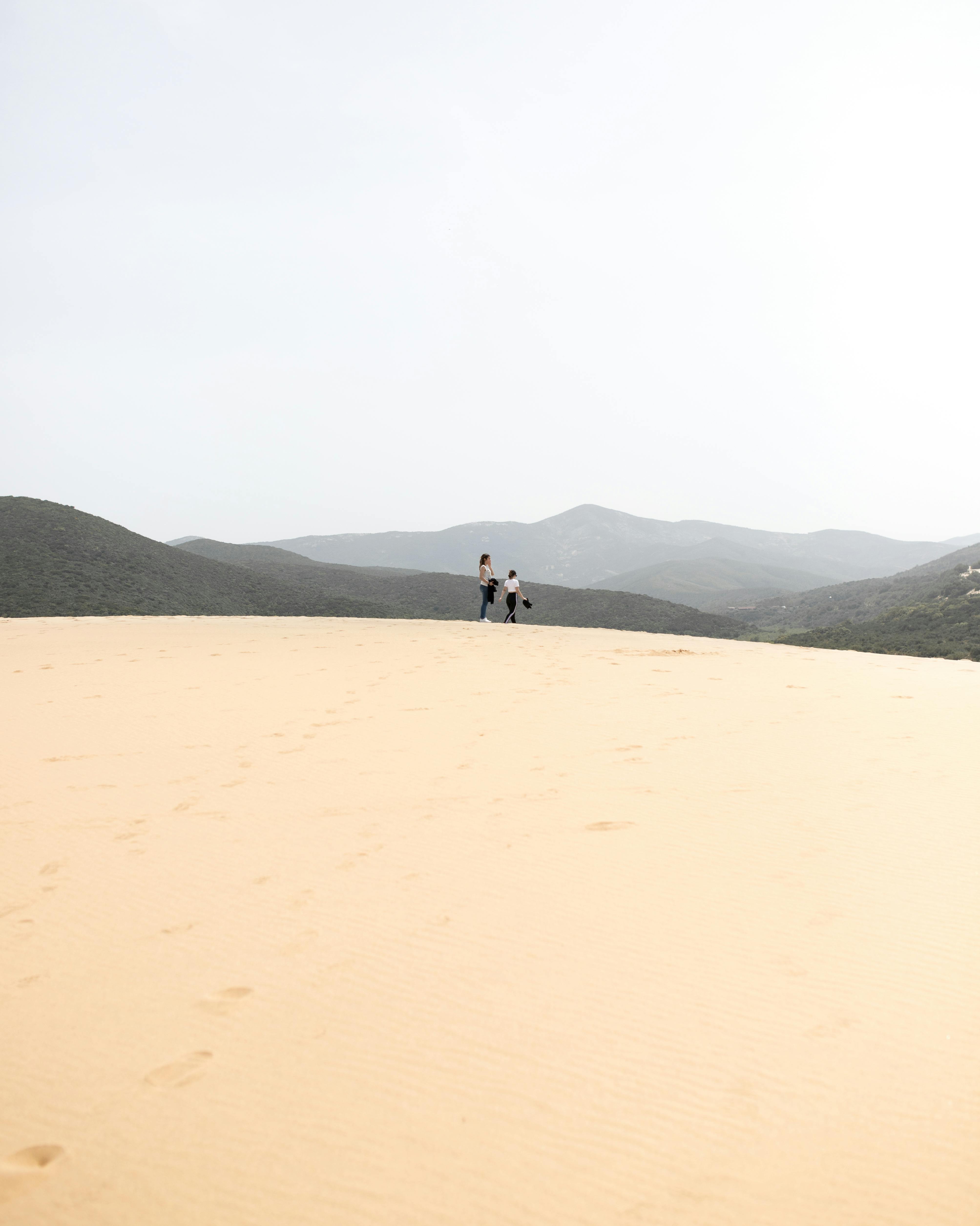 Two people traverse the expansive sandy dunes of Sardinia, Italy, under a bright sky.