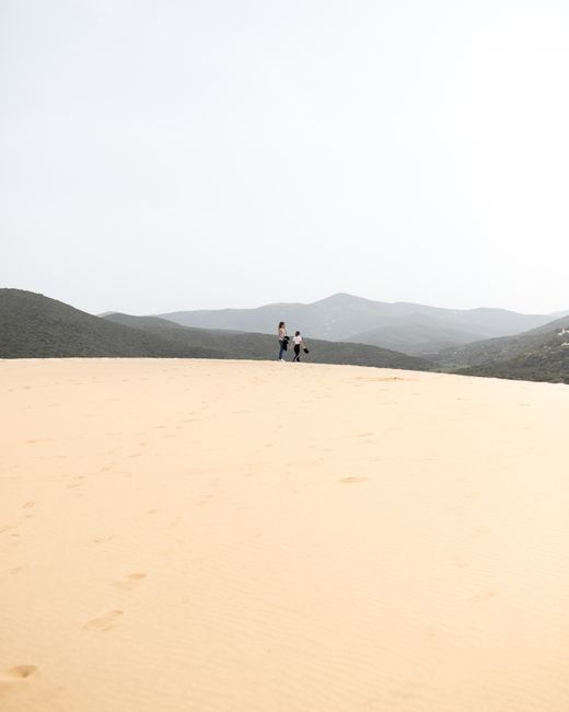Two people traverse the expansive sandy dunes of Sardinia, Italy, under a bright sky.