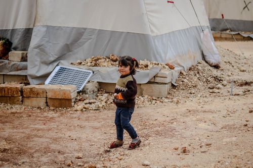 Free Full body of cheerful ethnic kid looking away while standing on ground near weathered tent in poor refugee camp in countryside Stock Photo