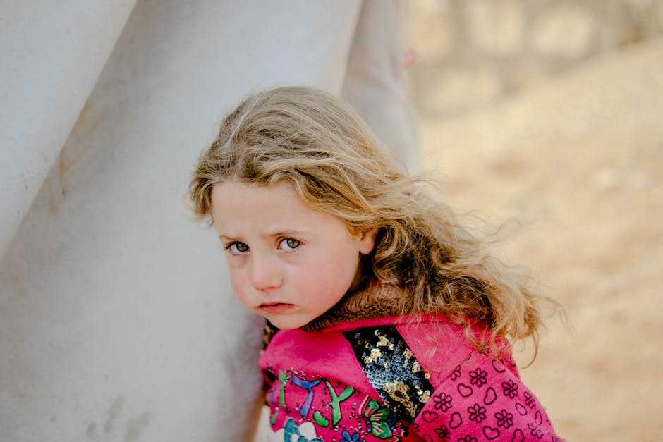 Unhappy little girl looking at camera while standing near weathered shelter in poor refugee camp on blurred background of countryside