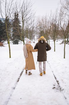 A couple in winter clothes walking on a snow-covered path in a park, captured from behind.